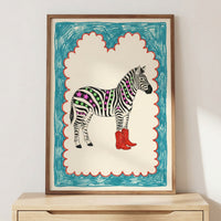 Framed artwork of a zebra with colorful, neon-like stripes filled with green lines and purple stars, wearing bright red cowboy boots. The playful drawing has a crayon-style blue and red scalloped border and is leaning on a wooden cabinet against a white wall.