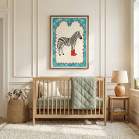 Framed zebra art featuring red cowboy boots and colorful striped stars hangs above a light wood crib in a softly lit nursery. The cozy room includes a sage green quilt, woven basket of stuffed animals, and a wooden side table with a lamp and books.