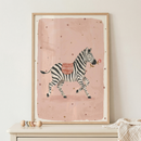 A whimsical illustration of a zebra is presented in a light-colored wooden frame. The zebra, depicted with its characteristic black and white stripes, is shown in motion, with one front leg lifted. It carries a small red and white checkered blanket on its back and holds a delicate pink flower in its mouth. The animal is set against a soft pink, rectangular background with an artistic texture, which is itself surrounded by a wide off-white border.