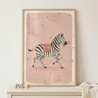 A whimsical illustration of a zebra is presented in a light-colored wooden frame. The zebra, depicted with its characteristic black and white stripes, is shown in motion, with one front leg lifted. It carries a small red and white checkered blanket on its back and holds a delicate pink flower in its mouth. The animal is set against a soft pink, rectangular background with an artistic texture, which is itself surrounded by a wide off-white border.