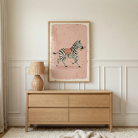 A vertical light wood-framed artwork hangs on a white paneled wall above a natural wood dresser. The artwork features an illustration of a black and white striped zebra trotting across a soft pink background dotted with small stars. The zebra wears a red and white checkered blanket on its back and holds a tiny pink flower in its mouth. On the four-drawer dresser sits a small lamp with a matching woven wicker base and lampshade.