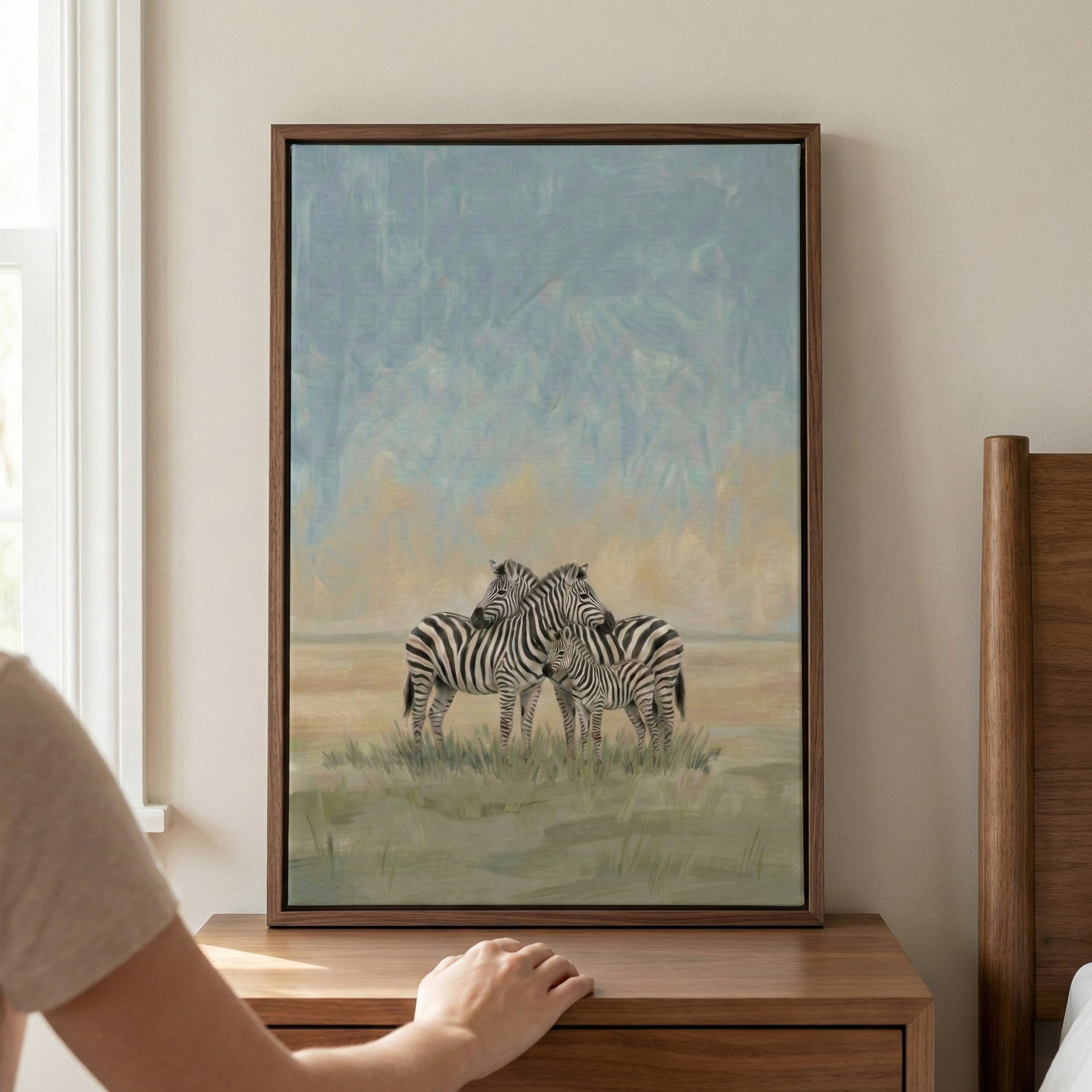 This framed wall art displays a serene scene of three zebras in an open field. Two adult zebras stand close together, with a small foal between them. The artwork features soft, muted colors and textured strokes, depicting the animals against a hazy sky of pale blue and yellow. The piece is held in a dark wooden frame and sits on a wooden dresser. A hand reaches toward the base of the frame, and a bedpost is visible on the right, indicating a domestic setting.