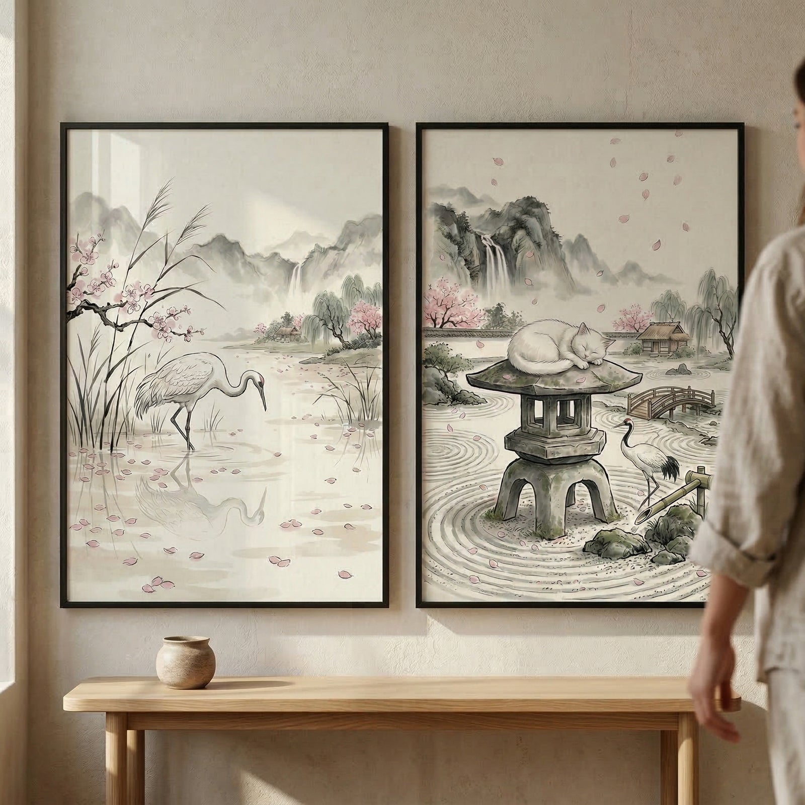 Two framed vertical artworks hang side-by-side on a light-colored wall above a simple wooden console table. The artwork on the left depicts a crane standing in a calm body of water with scattered pink flower petals. In the background are misty mountains and a waterfall. The artwork on the right shows a peaceful garden scene with a stone lantern.